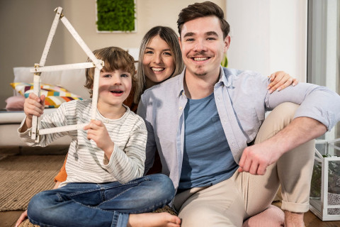 Wir unterstützen Sie schon bei der Suche nach Ihr Traumimmobilie. Vater, Mutter, Sohn sitzen im Wohnzimmer. Sohn hält Haus aus einem Zollstock gefaltet in der Hand.