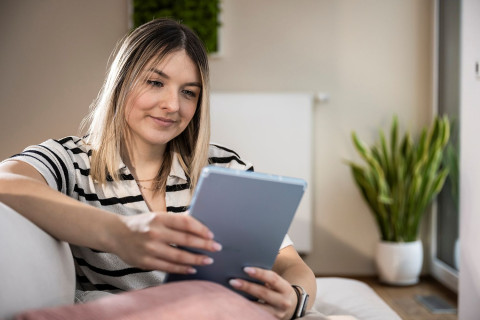 Sobald Sie in unserer Interessentendatenbank vertreten sind, lassen Ihnen unsere Immobilienmakler:innen vor Ort Angebote zukommen. Frau sitzt auf Sofa und schaut auf ihr Tablet.