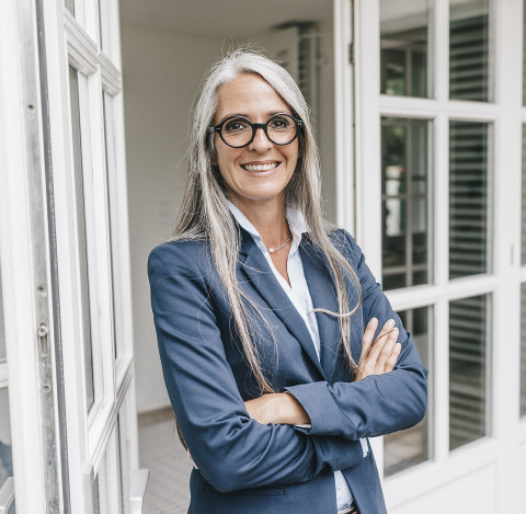 Immobilienmakler:innen unterstützen bereits bei der Vorbereitung des Verkaufs Maklerin mit grauen Haaren lächelnd lehnend an Terassentür