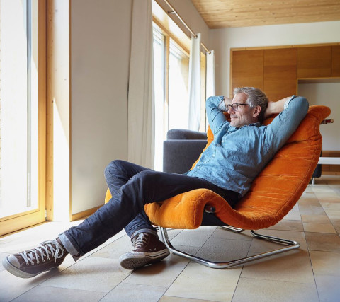 Die Aufgaben übernimmt der Makler und als Verkäufer können Sie sich zurücklehnen. Mann sitzt bequem und zurück gelehnt im Lesesessel. Die Arme hinter dem Kopf verschränkt.