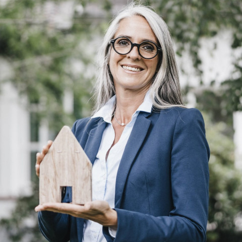 Termine, Besichtigungen und Vermarktung sind Aufgaben der Immobilienmakler:innen Maklerin präsentiert Holzhaus in den Händen.